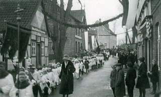 Kommunionkinder ziehen am Hochzeitshaus (l.) vorbei. Maria Welschof, Lehrerin an der Brinkschule, begleitet sie. Neben dem Hochzeitshaus stand Peters Eiche, dahinter das Gasthaus „Kösters" und die Jungen- oder Kirchschule. Das Gebäude rechts ist die heutige Tor-Deele. Die Aufnahme ist wohl aus dem Jahr 1938. - Fotosammlung Potthoff Kommunionkinder ziehen am Hochzeitshaus (l.) vorbei. Maria Welschof, Lehrerin an der Brinkschule, begleitet sie. Neben dem Hochzeitshaus stand Peters Eiche, dahinter das Gasthaus „Kösters" und die Jungen- oder Kirchschule. Das Gebäude rechts ist die heutige Tor-Deele. Die Aufnahme ist wohl aus dem Jahr 1938. - Fotosammlung Potthoff