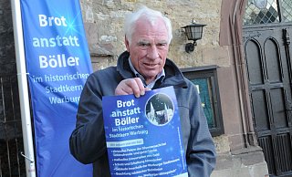 An einem Fahnenmast auf dem Brüderkichhof zwischen den Städten weht die neue Fahne der Aktion "Brott anstatt Böller". Mitinitiator Christian Voß präsentiert das Plakat, das aufmerksam machen soll. - Hubert Rösel An einem Fahnenmast auf dem Brüderkichhof zwischen den Städten weht die neue Fahne der Aktion "Brott anstatt Böller". Mitinitiator Christian Voß präsentiert das Plakat, das aufmerksam machen soll. - Hubert Rösel