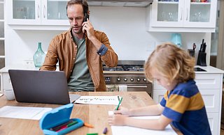 Ein Vater arbeitet zu Hause mit Smartphone und Laptop, während sein Sohn Schularbeiten macht. - picture alliance / Zoonar Ein Vater arbeitet zu Hause mit Smartphone und Laptop, während sein Sohn Schularbeiten macht. - picture alliance / Zoonar