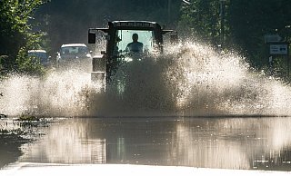 Ein Traktor fährt über eine Straße, die durch die starken Regenfälle unter Wasser steht. Im Juli und August waren die eigentliche ersehnten Niederschläge gebietsweise auch zu viel des Guten. - picture alliance/dpa Ein Traktor fährt über eine Straße, die durch die starken Regenfälle unter Wasser steht. Im Juli und August waren die eigentliche ersehnten Niederschläge gebietsweise auch zu viel des Guten. - picture alliance/dpa