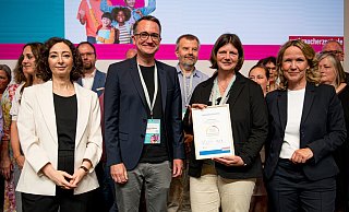 Bundesministerin Steffi Lemke (r.) und VZBV-Vorständin Ramona Pop (l.) zeichnen das Goerdeler-Gymnasium aus. Die Lehrkräfte Silke Michels und Sebastian Niggemann (Mitte) nehmen in Berlin als Mitglieder der Steuergruppe Urkunde und Schulplakette entgegen. - VZBV/Fabian Sommer Bundesministerin Steffi Lemke (r.) und VZBV-Vorständin Ramona Pop (l.) zeichnen das Goerdeler-Gymnasium aus. Die Lehrkräfte Silke Michels und Sebastian Niggemann (Mitte) nehmen in Berlin als Mitglieder der Steuergruppe Urkunde und Schulplakette entgegen. - VZBV/Fabian Sommer
