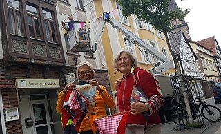 Eine von 70 Wimpelketten: Stadtwerke-Mitarbeiter Hannes Winter bringt vom Hubwagen aus die bunten Wimpel von Connie Tiemann (l.) und Monika Niemann über der Hauptstraße an. Rund 2.000 Exemplare hatten sie zusammen mit der Nähgruppe der "Zweiten Heimat" genäht. - Dieter Scholz Eine von 70 Wimpelketten: Stadtwerke-Mitarbeiter Hannes Winter bringt vom Hubwagen aus die bunten Wimpel von Connie Tiemann (l.) und Monika Niemann über der Hauptstraße an. Rund 2.000 Exemplare hatten sie zusammen mit der Nähgruppe der "Zweiten Heimat" genäht. - Dieter Scholz