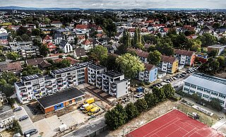 Links im Bild sind L-förmig die neuen Häuser zu sehen. Davor, orangefarben, der Treffpunkt mit seinen Beratungsstellen. Das aktuell schwarze Dach wird noch begrünt. Darauf folgen die Straße entlang drei Bestandshäuser der Genossenschaft sowie die Kita. Träger ist bei letzterer die AWO. - Gerald Dunkel Links im Bild sind L-förmig die neuen Häuser zu sehen. Davor, orangefarben, der Treffpunkt mit seinen Beratungsstellen. Das aktuell schwarze Dach wird noch begrünt. Darauf folgen die Straße entlang drei Bestandshäuser der Genossenschaft sowie die Kita. Träger ist bei letzterer die AWO. - Gerald Dunkel
