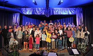 Das Bild zeigt den Chor der PaderVoices auf der Bühne gemeinsam mit ihren fröhlichsten Fans, den Flüchtlingskindern. Einige Kinder halten Prilblumen hoch, die zum Saalschmuck im Stil der Siebziger gehörten - xxxxxxxx Das Bild zeigt den Chor der PaderVoices auf der Bühne gemeinsam mit ihren fröhlichsten Fans, den Flüchtlingskindern. Einige Kinder halten Prilblumen hoch, die zum Saalschmuck im Stil der Siebziger gehörten - xxxxxxxx